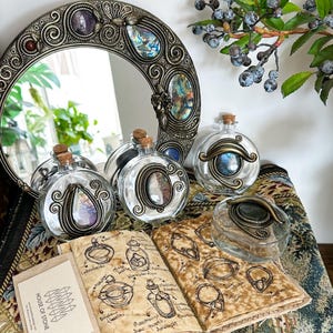 May include: A collection of decorative glass bottles with cork stoppers and ornate metal frames. The bottles are arranged with a round mirror and a journal with sketches. The mirror and bottles feature colorful gemstones.