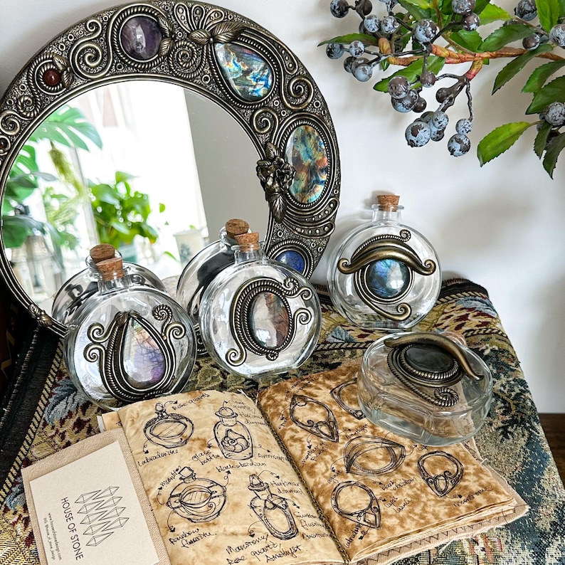 May include: A collection of decorative glass bottles with cork stoppers and ornate metal frames. The bottles are arranged with a round mirror and a journal with sketches. The mirror and bottles feature colorful gemstones.