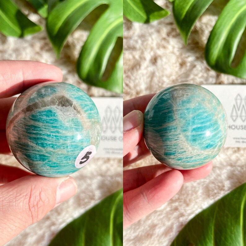 May include: A smooth, polished green and white amazonite sphere, approximately 2 inches in diameter. The sphere has a natural, striated pattern and is held in a hand.