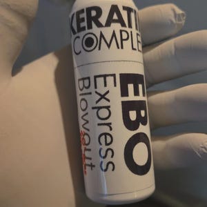 May include: A white bottle with a black cap and black text. The bottle reads "KERATIN COMPLEX" and "EBO Express Blowout". The bottle is held by a gloved hand.