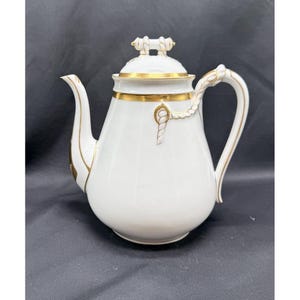 May include: A white porcelain teapot featuring gold trim accents on the lid, neck, and spout. The handle and lid finial are designed with a sculpted rope motif. The body has a smooth, rounded shape with a curved spout and a sturdy handle.