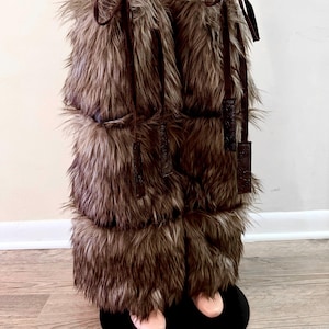 May include: Brown faux fur leg warmers with leather-like ties. The leg warmers are made of long, shaggy faux fur and are designed to be worn over the lower legs. The ties are a darker brown and add a decorative element to the warmers.