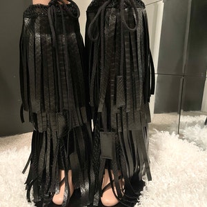 May include: Black faux leather leg wraps with fringe detailing. The wraps feature a drawstring closure at the top and are designed to be worn over the lower legs. The fringe hangs down in multiple layers, creating a stylish and edgy look.