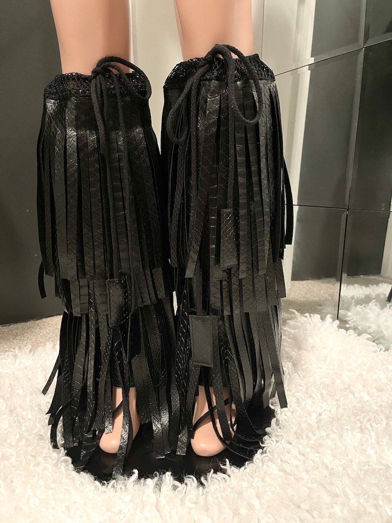 May include: Black faux leather leg wraps with fringe detailing. The wraps feature a drawstring closure at the top and are designed to be worn over the lower legs. The fringe hangs down in multiple layers, creating a stylish and edgy look.