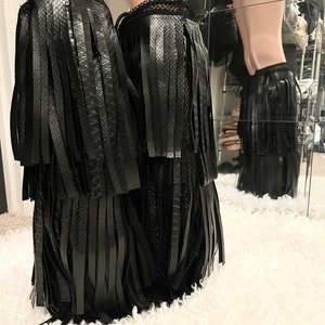 May include: Black faux leather leg wraps with fringe detailing. The wraps feature a textured, snake-like pattern and a lace trim at the top. The wraps are displayed on a mannequin leg.
