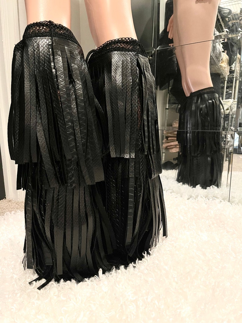 May include: Black faux leather leg wraps with fringe detailing. The wraps feature a textured, snake-like pattern and a lace trim at the top. The wraps are displayed on a mannequin leg.