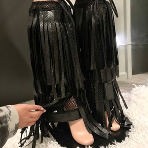 May include: Black faux leather leg wraps with long fringe detailing. The wraps feature a textured snake-like pattern and are secured with ties at the top. The design includes a mesh panel near the ankle and a strap around the foot.