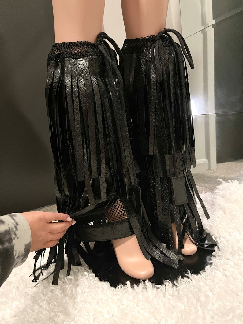 May include: Black faux leather leg wraps with long fringe detailing. The wraps feature a textured snake-like pattern and are secured with ties at the top. The design includes a mesh panel near the ankle and a strap around the foot.