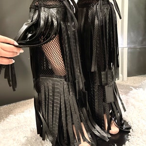 May include: Black faux leather leg wraps with fringe detailing. The wraps feature a mesh upper and are tied with black cords. The design includes a textured, snake-like pattern on the faux leather.