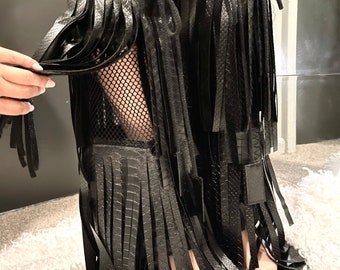 May include: Black faux leather leg wraps with fringe detailing. The wraps feature a mesh upper and are tied with black cords. The design includes a textured, snake-like pattern on the faux leather.