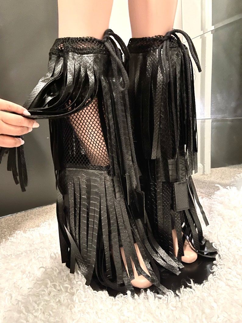 May include: Black faux leather leg wraps with fringe detailing. The wraps feature a mesh upper and are tied with black cords. The design includes a textured, snake-like pattern on the faux leather.
