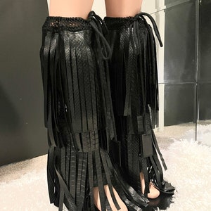 May include: Black fringed leg warmers. These leg warmers feature a textured, snake-like pattern and are adorned with long, vertical fringe. The top of the leg warmers has a crocheted trim and a drawstring closure. The leg warmers are shown on a mannequin.