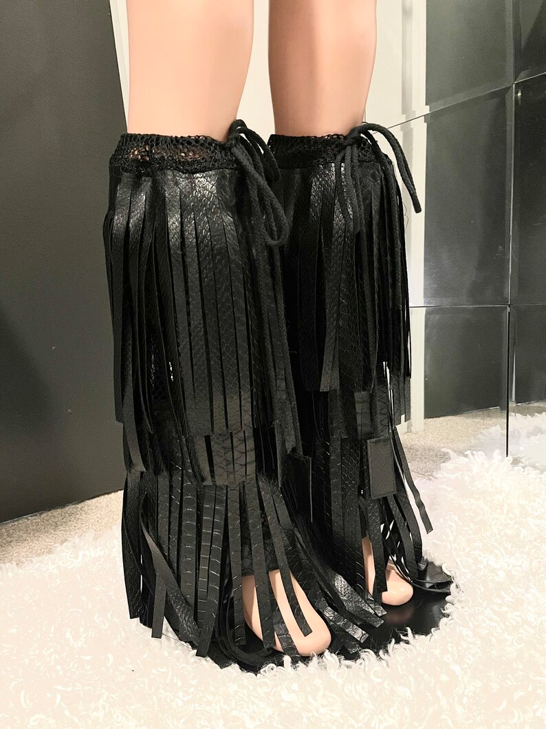 May include: Black fringed leg warmers. These leg warmers feature a textured, snake-like pattern and are adorned with long, vertical fringe. The top of the leg warmers has a crocheted trim and a drawstring closure. The leg warmers are shown on a mannequin.