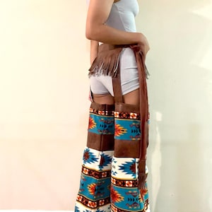 May include: Brown leather chaps with a fringed waist detail and a colorful southwestern pattern on the lower legs. The pattern features turquoise, orange, and white geometric designs. The chaps have a high-heeled boot base.
