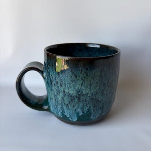 May include: A ceramic mug with a dark blue and teal glaze. The mug has a rounded body, a large handle, and a glossy finish. The glaze has a textured, dripped appearance, with darker hues at the top and lighter shades towards the bottom.