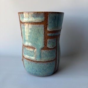 May include: A handmade ceramic tumbler with a unique design. The exterior features a light blue glaze with a brown, textured grid pattern. The interior is a darker brown. The tumbler has a slightly irregular shape.