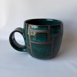 May include: A teal ceramic mug with a dark handle. The mug features a geometric pattern in a darker teal color. The interior of the mug is dark. The mug is on a white background.