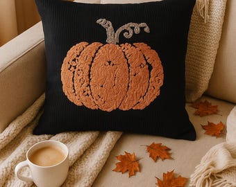 May include: Black square pillow with an embroidered orange pumpkin design. The pillow is on a beige couch with a cream-colored blanket and a cup of coffee. Autumn leaves are scattered around.