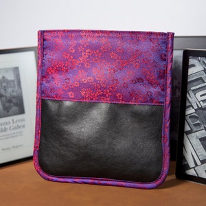 Könnte beinhalten: Eine E-Reader-Hülle aus violett-rotem Stoff mit floralem Muster und einer schwarzen Lederklappe. Die Hülle ist geschlossen und liegt auf einer braunen Oberfläche.