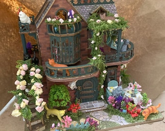 May include: A detailed miniature cat house with a brick facade, balconies, and a garden. The house features multiple levels, with small cat figurines on the balconies and in the garden. The house is decorated with flowers and greenery, creating a whimsical scene.