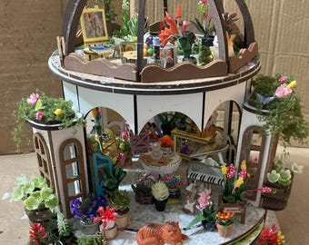 May include: A miniature wooden greenhouse with a domed roof, featuring detailed interior scenes. The structure is painted white and brown, with miniature plants, furniture, and figurines. The scene includes a piano, tables, and a sleeping cat figurine.