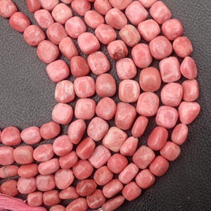 May include: Strands of pink rhodochrosite gemstone beads. The beads are a mix of rounded and slightly irregular shapes, with a polished, smooth surface. The color varies from light to medium pink, with some beads showing subtle variations in tone. The beads are strung together.