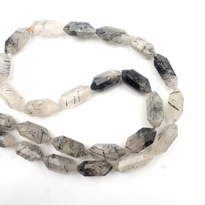 Puede incluir: Un collar de cuentas alargadas y facetadas. Las cuentas son transparentes con inclusiones negras, creando un efecto moteado. Las formas geométricas y los tonos naturales crean una estética terrosa.