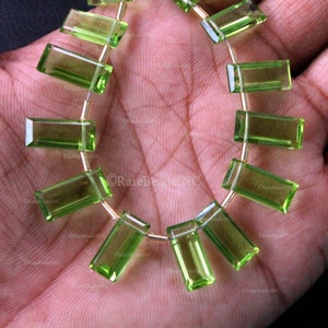 May include: A strand of translucent, light green, rectangular-cut gemstones arranged in a curved formation. Each individual stone features faceted edges and is strung together with thin, metallic wire, creating a uniform, repeating pattern.