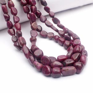 May include: Close-up of strands of polished, deep red ruby gemstone beads. The beads are smooth and rounded, varying in size. The gemstones are strung together, suitable for a necklace or bracelet. The background is white.