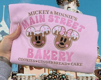 May include: A light pink sweatshirt featuring an embroidered design of Mickey and Minnie Mouse gingerbread cookies. The text reads "Mickey & Minnie's Main Street Bakery" with "Cookies Gingerbread Cake" below. The background shows a castle.