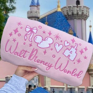 Puede incluir: Un art&iacute;culo rectangular rosa con el texto bordado "Walt Disney World" y varios dise&ntilde;os bordados en blanco y rosa, incluyendo orejas de Mickey, corazones y estrellas. El fondo presenta un castillo con torres azules.