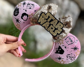 Könnte beinhalten: Rosa Micky-Maus-Ohren mit Star Wars-Motiven. Die Ohren haben eine goldene Pailletten-Schleife und ein goldglitzerndes "Star Wars"-Emblem. Die Ohren zeigen verschiedene Star Wars-Charakterillustrationen und goldene Sternakzente.
