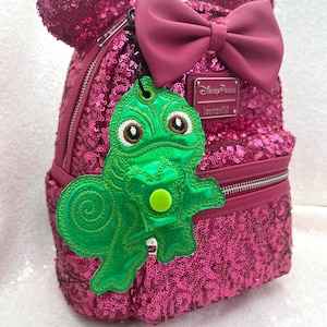 May include: Pink sequined Minnie Mouse backpack with a pink bow and a green chameleon keychain. The keychain has the text "Disney Parks" and "Loungefly" on it.