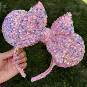 May include: Pink sequined mouse ear headband with a large bow. The ears and bow are covered in iridescent sequins that catch the light. The headband is a light pink color.