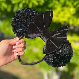 May include: Black sequined mouse ear headband with a large velvet bow. The ears are covered in small, shiny black sequins. The bow is made of a soft, black velvet material. The headband is held by a hand.