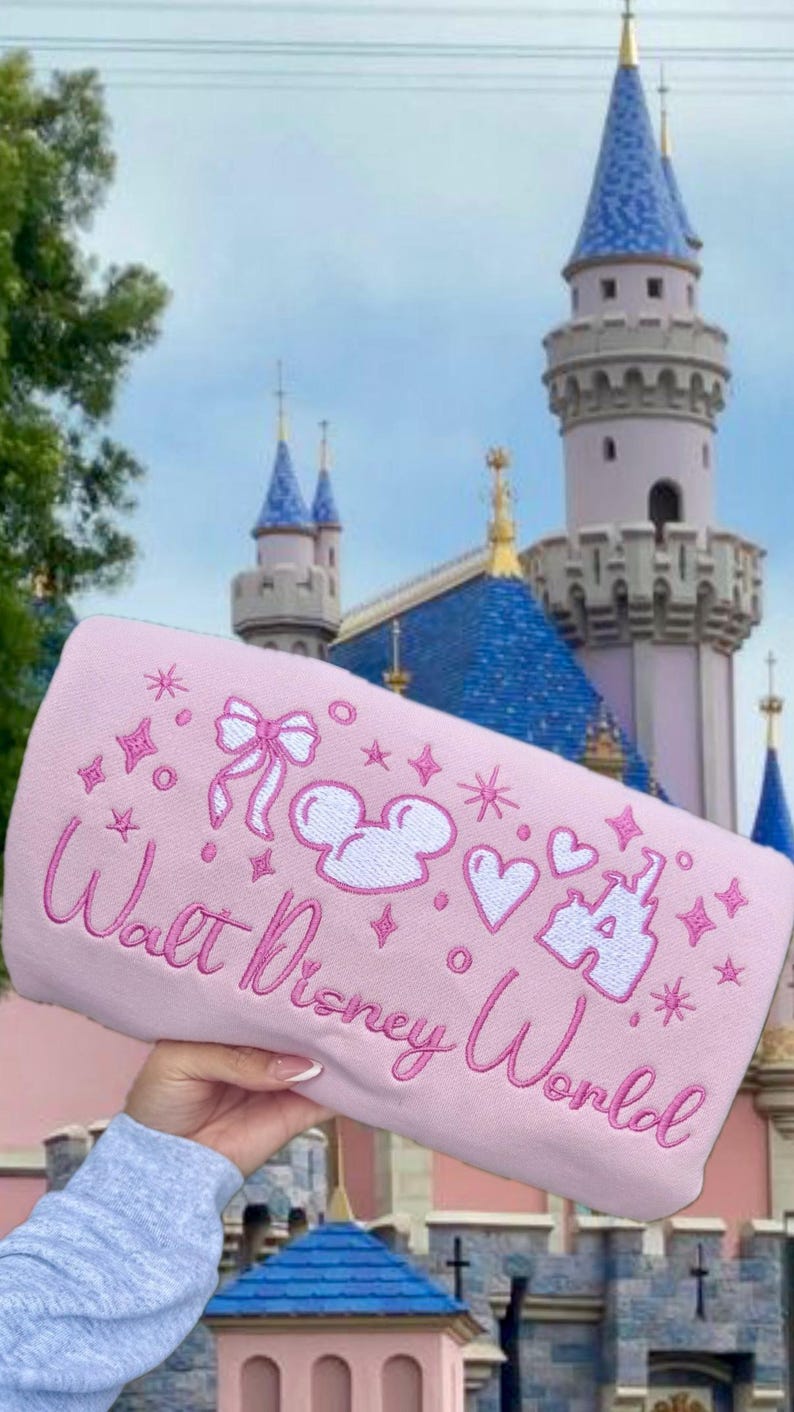 Puede incluir: Un art&iacute;culo rectangular rosa con el texto bordado "Walt Disney World" y varios dise&ntilde;os bordados en blanco y rosa, incluyendo orejas de Mickey, corazones y estrellas. El fondo presenta un castillo con torres azules.