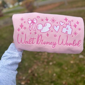 Puede incluir: Una sudadera rosa claro con gr&aacute;ficos blancos y rosas bordados. El dise&ntilde;o incluye un lazo, cabezas de Mickey Mouse, corazones, estrellas y un castillo. Las palabras "Walt Disney World" est&aacute;n bordadas en cursiva.