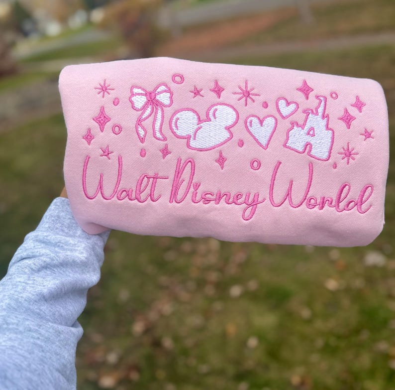 Puede incluir: Una sudadera rosa claro con gr&aacute;ficos blancos y rosas bordados. El dise&ntilde;o incluye un lazo, cabezas de Mickey Mouse, corazones, estrellas y un castillo. Las palabras "Walt Disney World" est&aacute;n bordadas en cursiva.