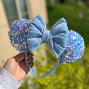 May include: Light blue sequined mouse ears with a matching velvet bow. The headband is light blue with a white stripe. The ears are covered in iridescent sequins.