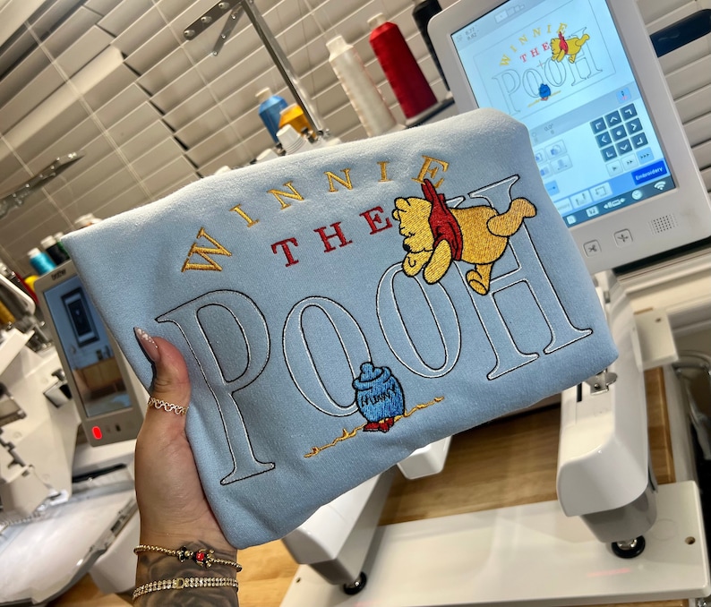 May include: A light blue sweatshirt with embroidered text that reads "Winnie the Pooh" in gold, red, and white. The embroidery also includes a cartoon image of Winnie the Pooh holding a jar of honey.