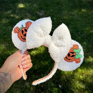 May include: White plush headband with a large bow and two round ears. Each ear features an embroidered orange pumpkin with a smiling face. The headband and bow are made of a soft, textured material, perfect for Halloween.