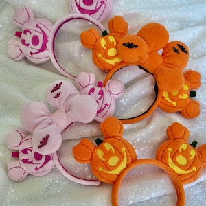 May include: Four headbands featuring plush Mickey Mouse-shaped pumpkin designs. Two headbands are orange with yellow pumpkin faces and green stems, while two are light pink with dark pink pumpkin faces and include large plush bows with bat icons.