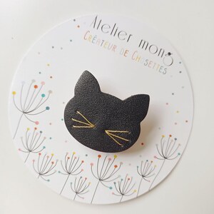 Op de afbeelding: Zwarte leren katten broche met gouden draad borduurwerk. De broche is op een witte achtergrond met een kleurrijk paardenbloem patroon. De tekst "Atelier Mono Créateur de Chosseties" is op de achtergrond gedrukt.