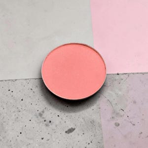 Puede incluir: Una paleta de maquillaje redonda de color coral con un borde de metal plateado. El colorete se encuentra sobre un fondo de superficies grises y rosas, creando una composición sencilla pero visualmente atractiva. El producto es probablemente un colorete o una sombra de ojos.