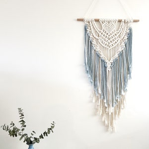 May include: A white and blue macrame wall hanging with a geometric pattern and fringe. The hanging is attached to a wooden dowel.