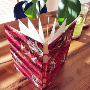 May include: A red and gold fabric-covered journal with a Japanese-inspired design of fish and waves. The journal is open to reveal blank pages.
