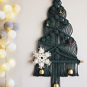 Pode incluir: Um pendurado de parede de árvore de Natal verde em macramê com um floco de neve branco e sinos dourados. A árvore é decorada com pequenos enfeites vermelhos e verdes.