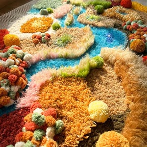 Könnte beinhalten: Ein farbenfroher, strukturierter Teppich mit Landschaftsdesign. Der Teppich zeigt einen blauen Fluss, umgeben von verschiedenen Texturen und Farben, darunter Orange, Grün, Rosa und Braun. Über die Oberfläche sind verschiedenfarbige Pompons verteilt.