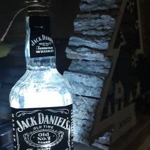 Peut inclure: Une bouteille en verre transparent avec une étiquette noire qui indique "Jack Daniel's Old No. 7 Brand Tennessee Sour Mash Whiskey 0.701 40% vol". La bouteille est éclairée de l'intérieur, créant une lueur blanche brillante.