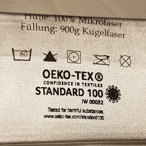 May include: A silver-coloured fabric care label featuring the text H&uuml;lle: 100% Mikrofaser, F&uuml;llung: 900g Kugelfaser, and OEKO-TEX Standard 100. The label includes five laundry care symbols, the OEKO-TEX logo, and the website www.oeko-tex.com/standard100.